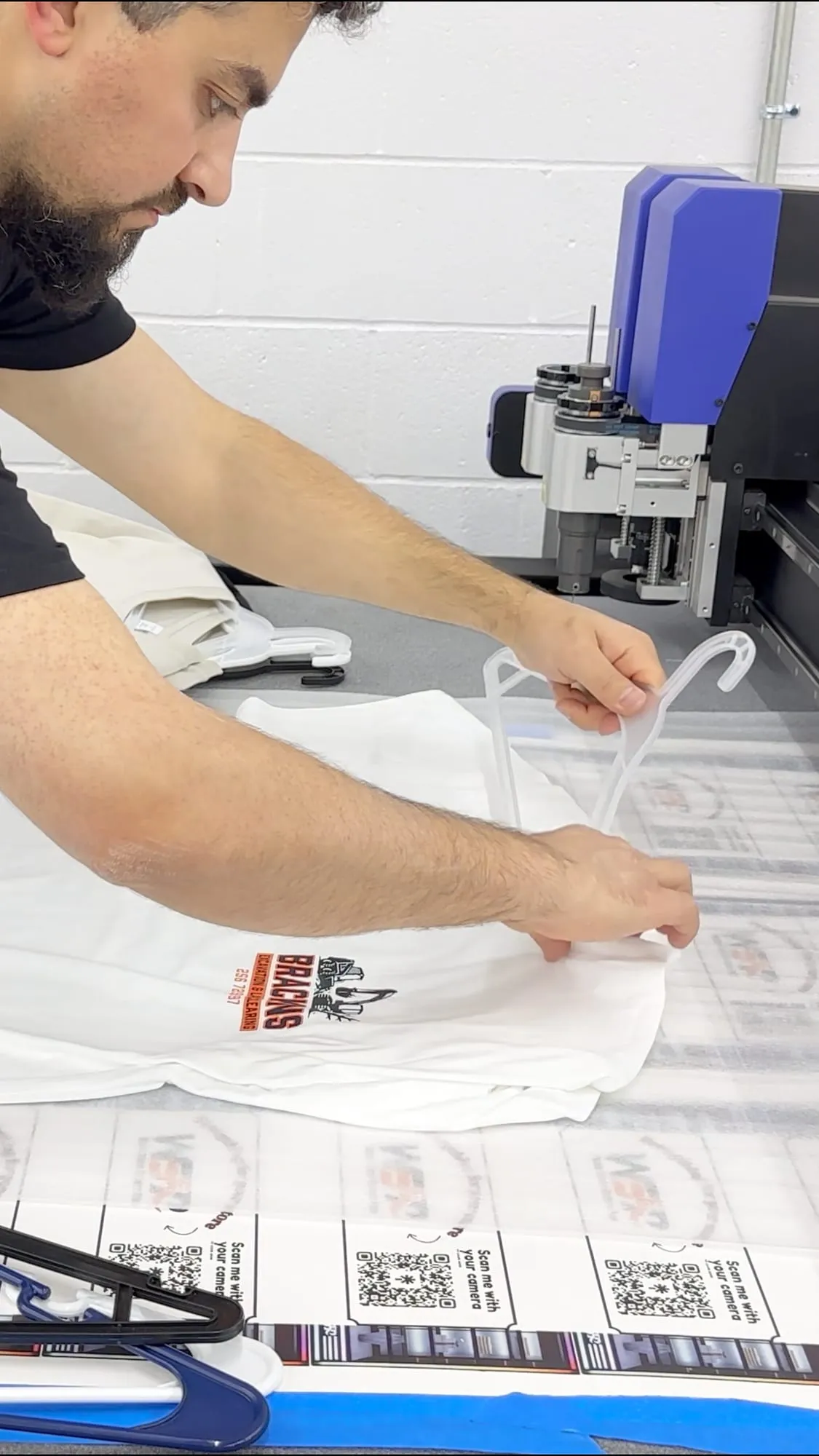 A person with a beard folds a white T-shirt with a small printed logo on a cutting table beside a large flatbed cutter, with a sheet of printed QR-code labels along the front edge of the table.