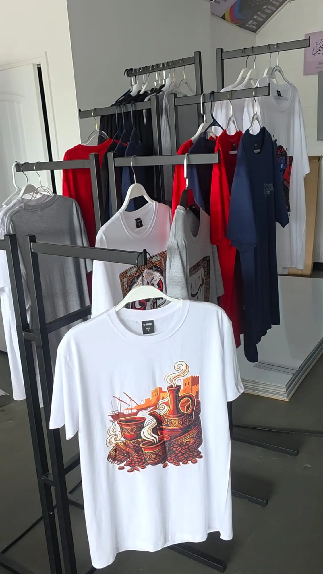 A white T-shirt printed with an illustration of steaming coffee pots, cups and coffee beans hangs on a black rack in front of red, navy, gray and white shirts, some with other prints.