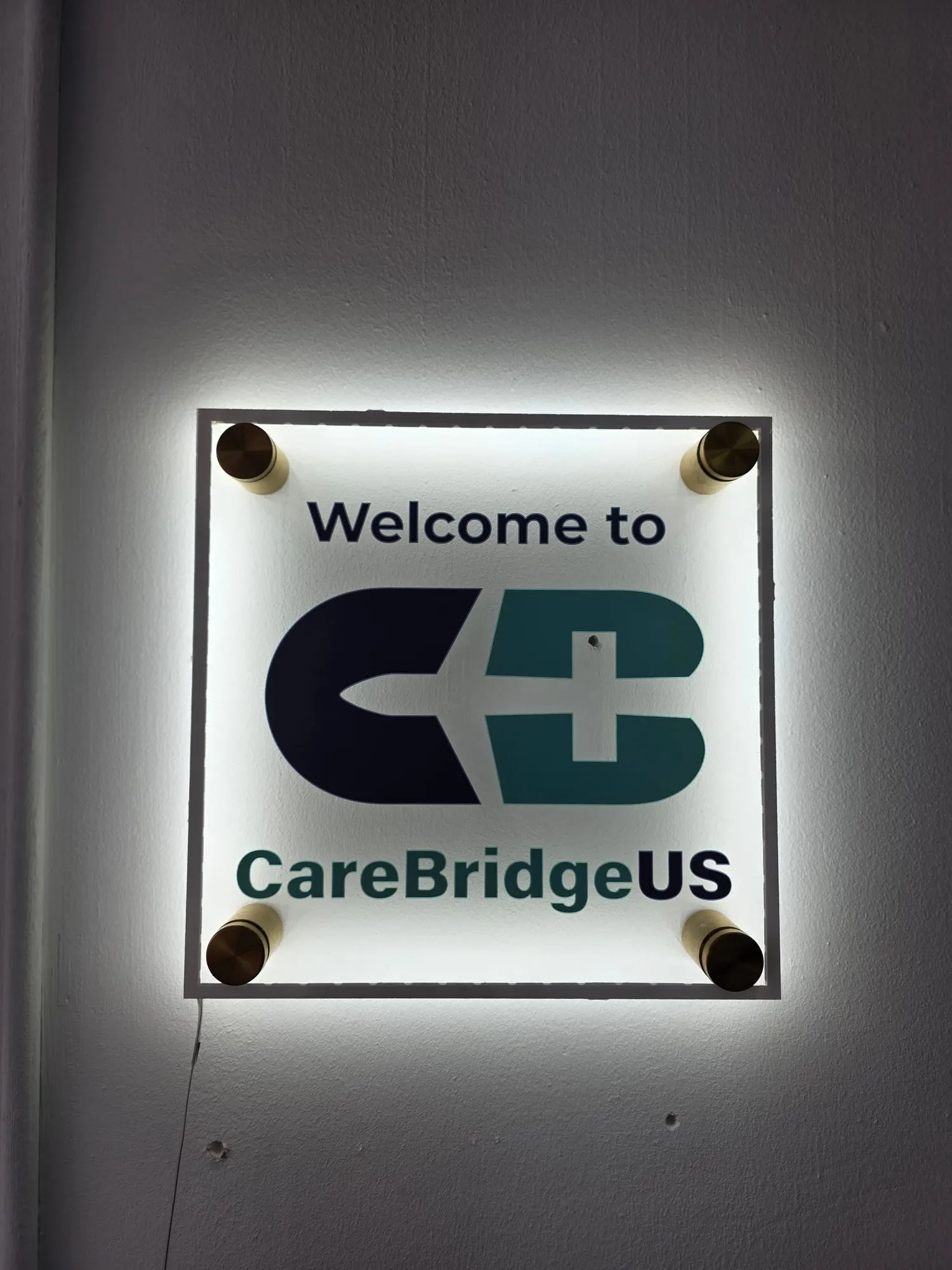 Square sign panel on a gray wall, lit from behind with white light, reading Welcome to above a two-tone logo and a name line, held off the wall by four gold-colored standoffs.