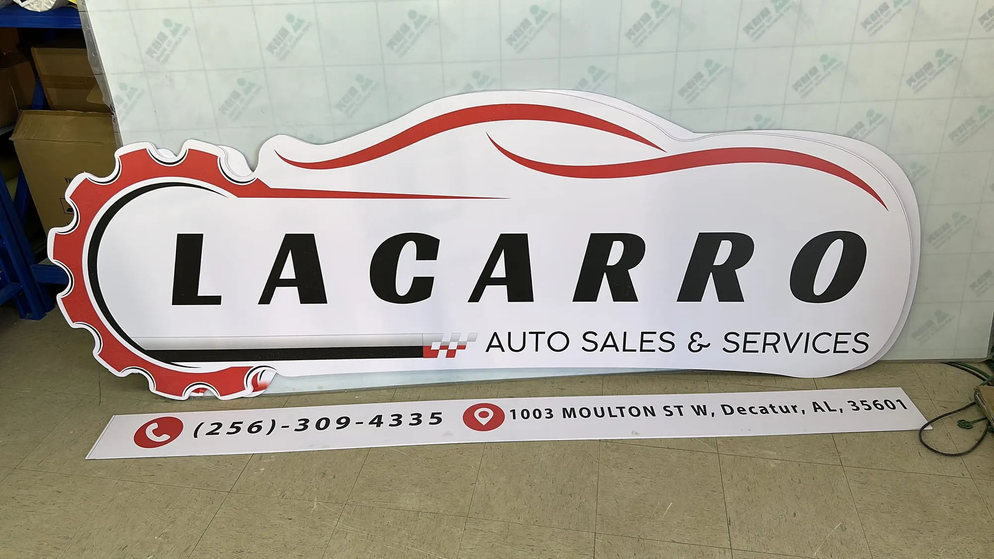 A white sign cut in a gear and swoosh outline lying flat on a tiled floor with red and black lettering, and a long strip sign on the floor in front of it.