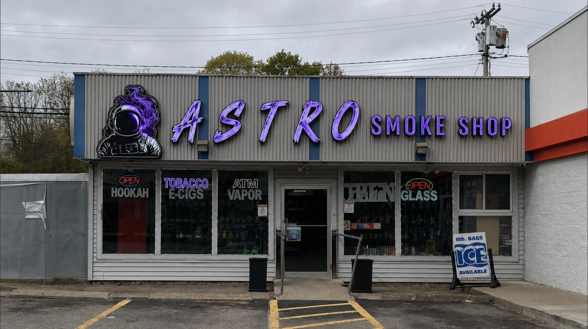 Storefront by day with purple lettering and an astronaut graphic on a gray sign band above windows with white lettering and small lit open signs.
