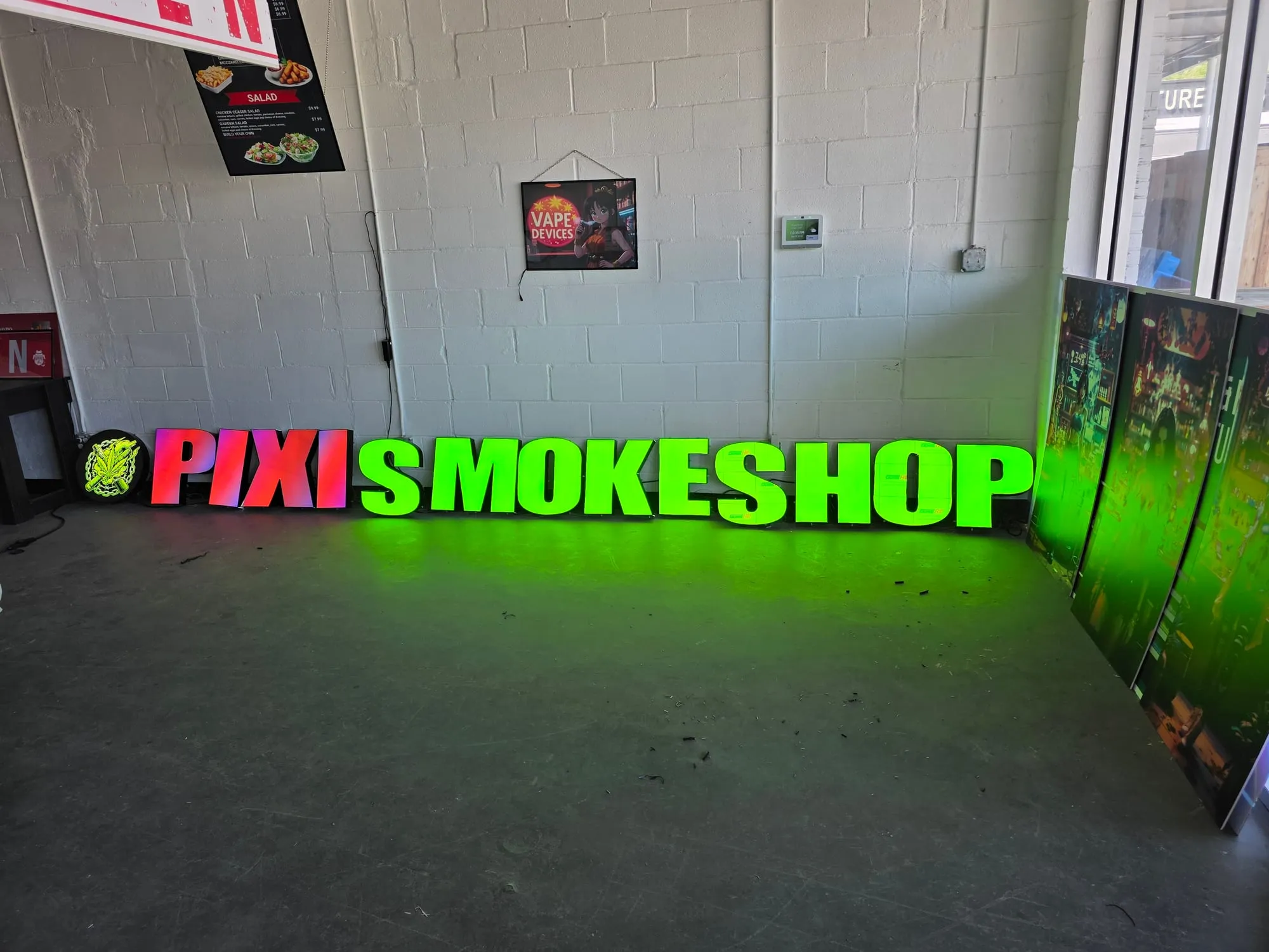 A row of large lit channel letters on a concrete floor in front of a white brick wall, the first four glowing pink and red and the rest bright green, with a round logo at the left end.