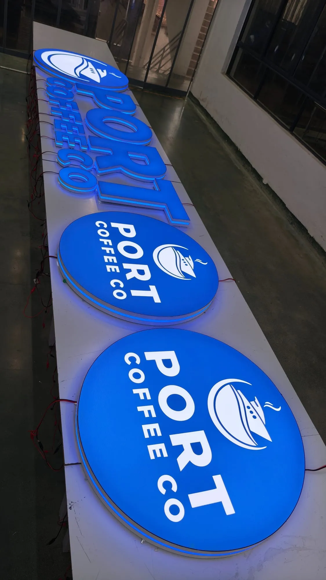 Two round signs and a set of dimensional letters lit bright blue on a long white worktable, each round face showing white lettering and a ship-and-cup logo.