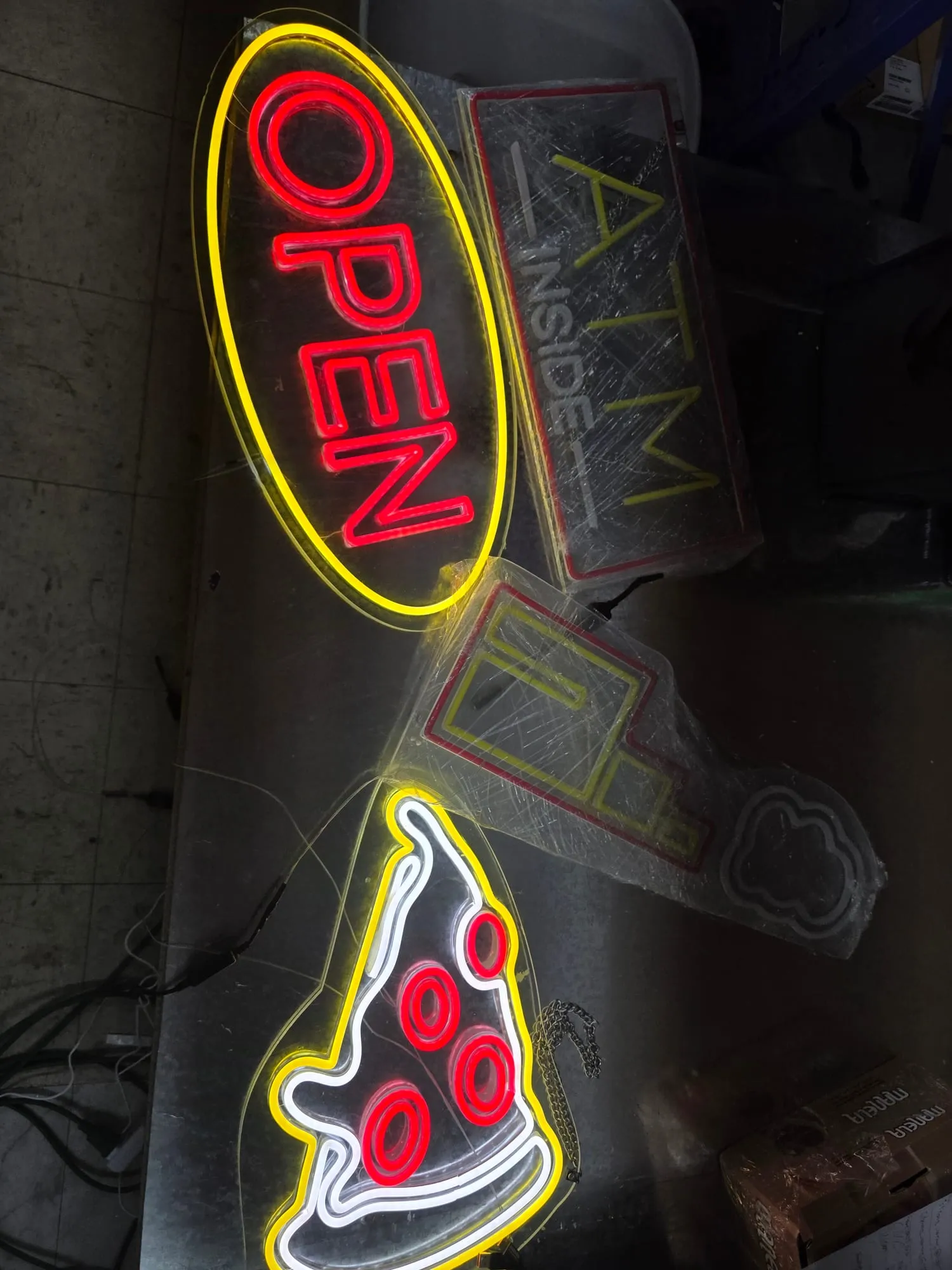 Several lit signs lying on a metal workbench: a red OPEN inside a yellow oval, an ATM INSIDE sign, and a pizza slice outlined in yellow, white and red.