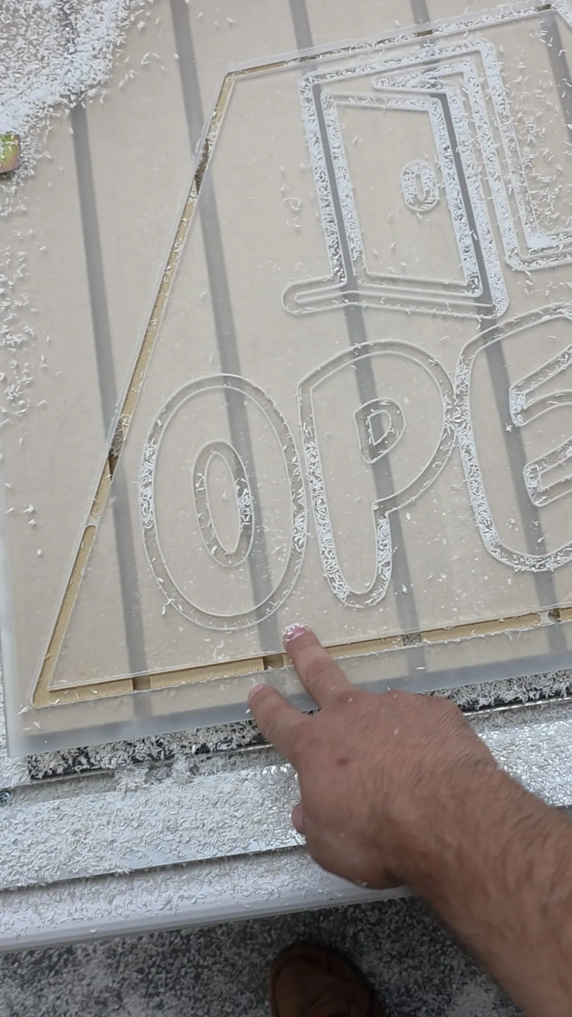 A hand points to the edge of a clear panel on a cutting table, with an OPEN outline and a second shape cut into it and white shavings around the cut lines.