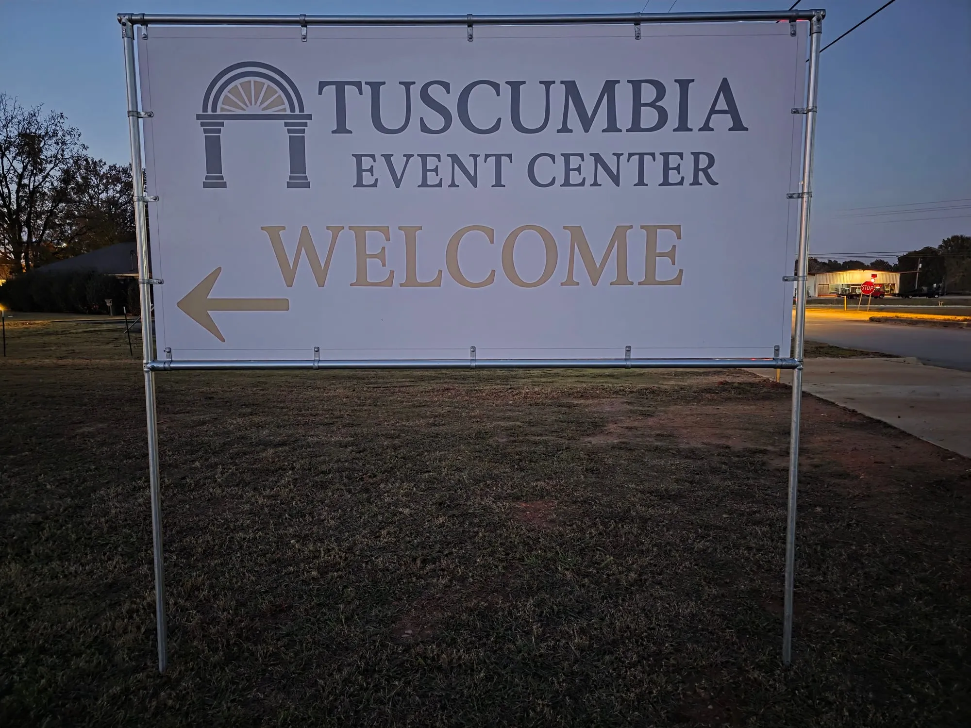 Printed banner on a silver pipe frame in a grass lawn at dusk, with an arch logo, an event center name, the word WELCOME and a left-pointing arrow.