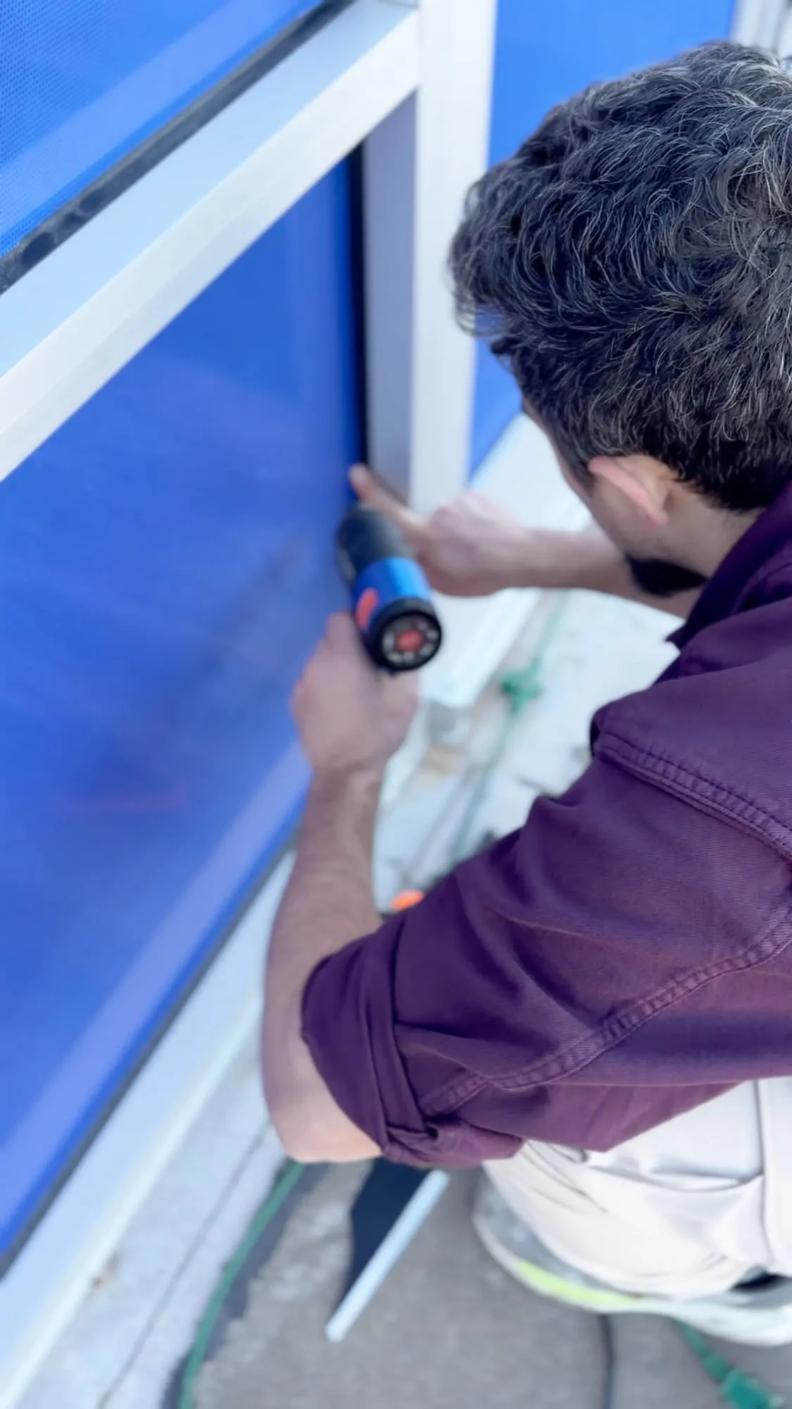 A person in a purple shirt crouches at a window frame and presses a heat gun against the blue printed window film.