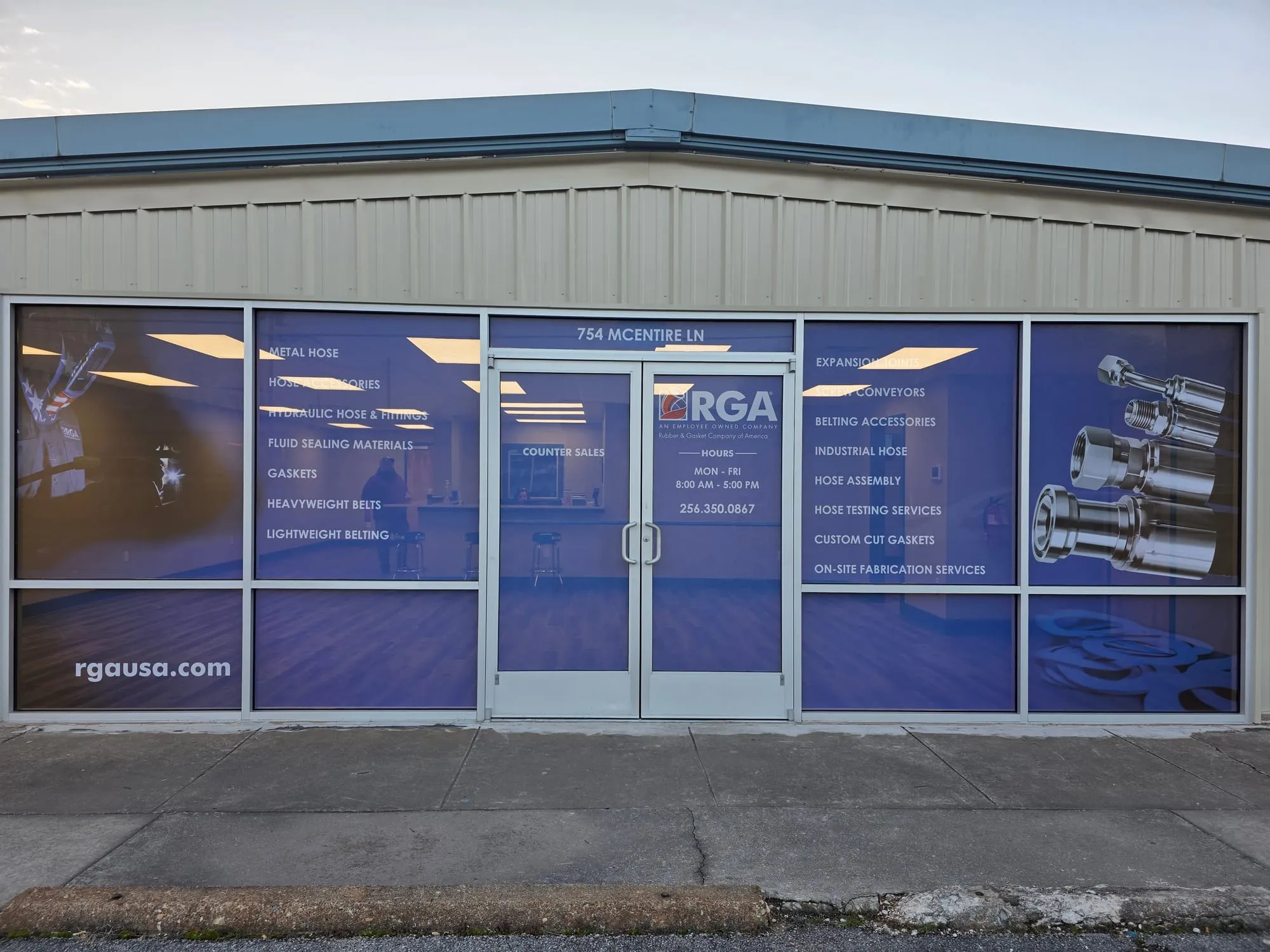Front of a tan metal building with blue-toned printed graphics on all the windows and the double glass doors, including a logo, business hours, a product list and photos of chrome fittings.