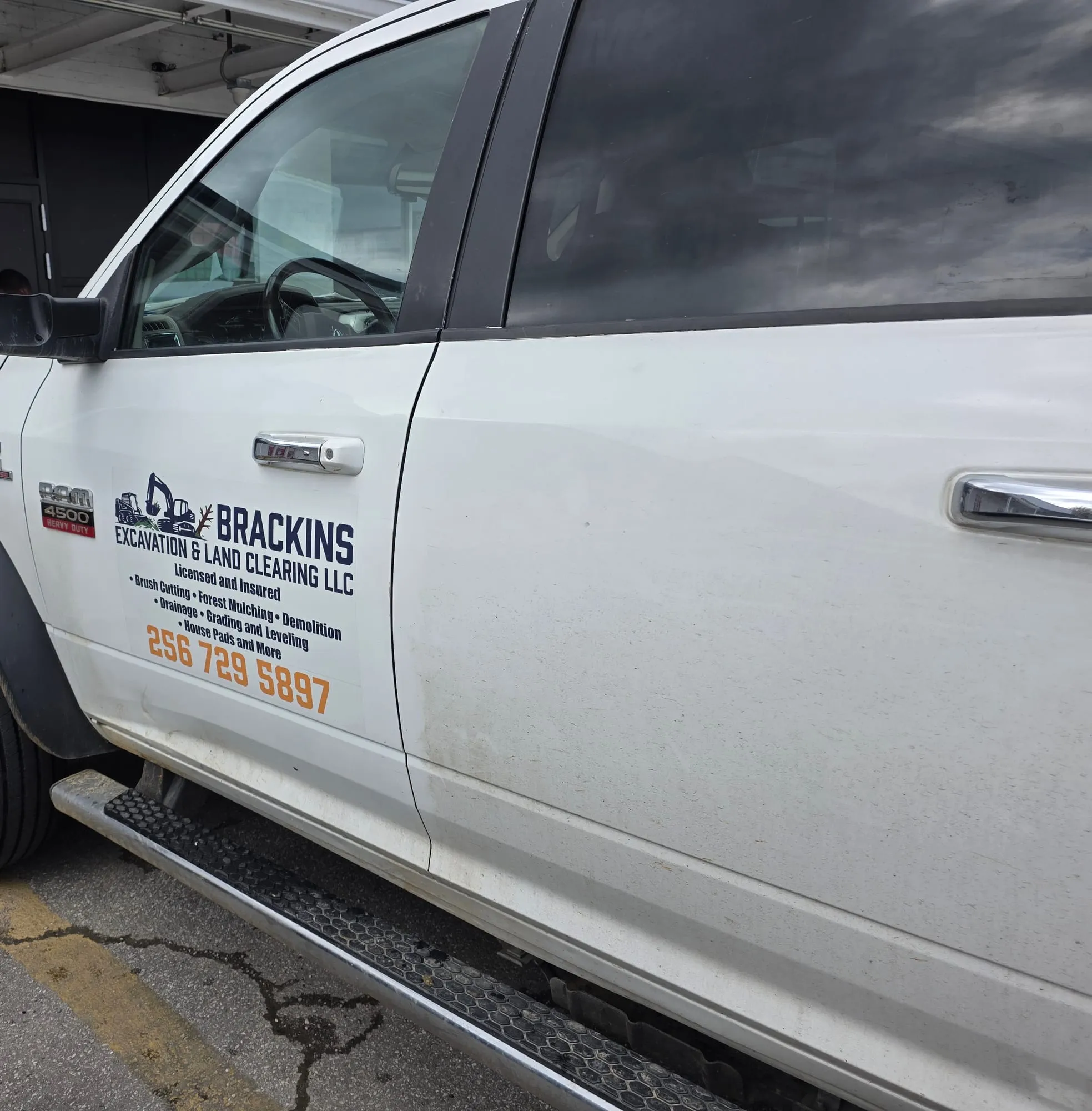 Front door of a white heavy-duty pickup with a printed graphic showing an excavator illustration, a company name, a list of services and a large orange phone number.